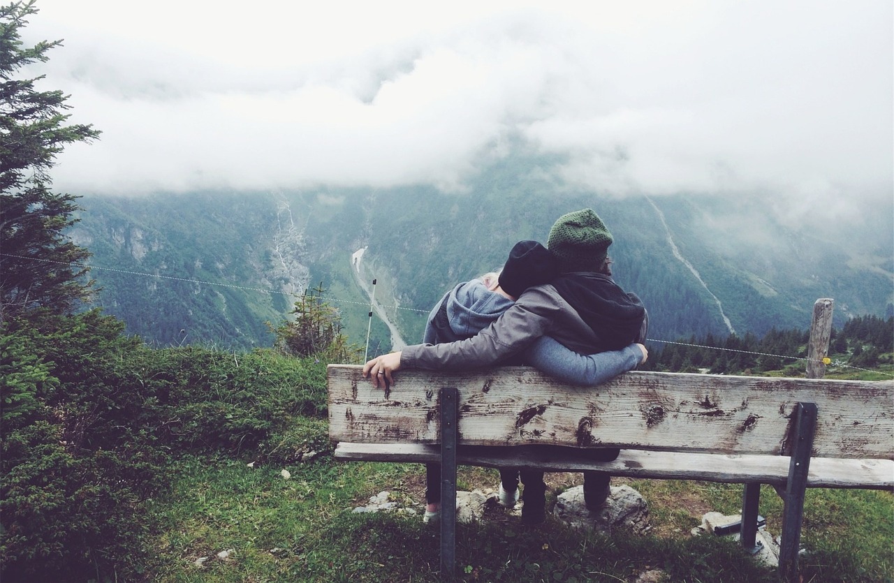 Junges Pärchen sitzt Arm in Arm auf einer Bank in den Bergen