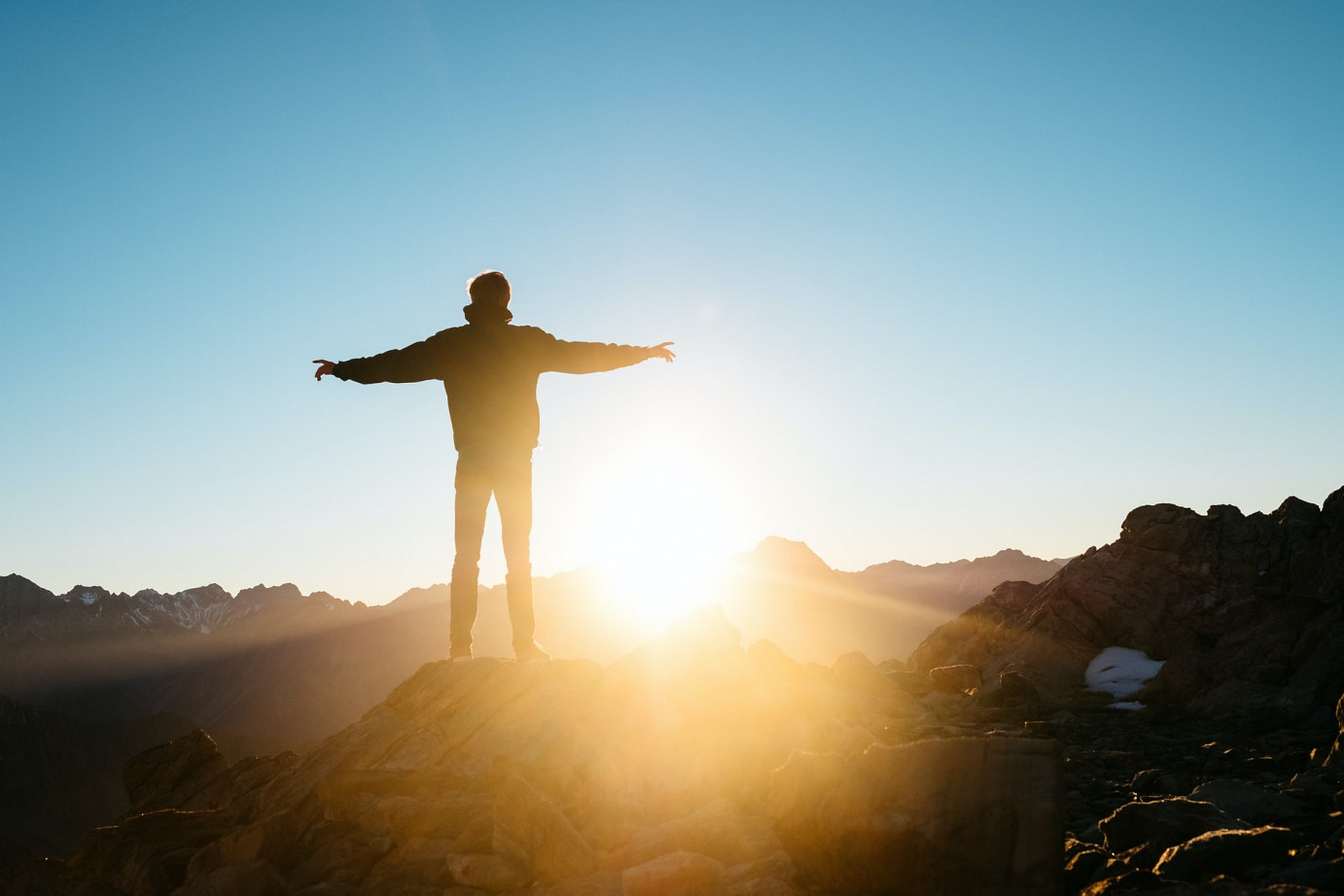 Mann steht auf einem Gipfel und blickt in den Sonnenuntergang